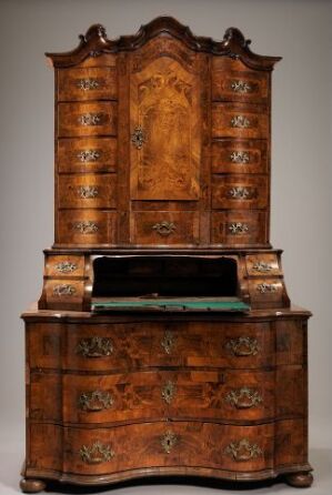  An elegant antique wooden bureau-cabinet with a serpentine front, standing against a gray backdrop. It has four drawers with ornate brass handles, a fold-down green felt-covered writing surface, and an arched two-door cabinet section with detailed wood grain panels and brass fittings.