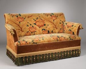  An antique-style sofa with rolled arms and a floral pattern featuring orange and rust flowers against a light tan background, complete with a dark green and beige tassel fringe along the base.