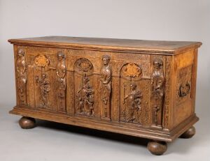  An antique wooden chest with intricate carvings featuring historical figures on its panels, standing on rounded feet, against a neutral background.