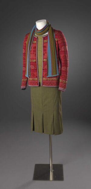  Vintage woman's outfit on a mannequin consisting of a long-sleeve cardigan with red and gold horizontal stripes, a light-collared blouse, and a muted olive green pleated knee-length skirt against a dark grey gradient background.