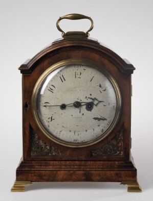  Antique mantel clock with arched wooden casing, Roman numeral clock face behind glass, brass carrying handle on top, and a color palette of deep wooden browns, muted brass, and weathered black.