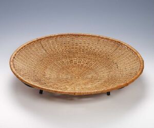  A shallow, handwoven basket with a natural tan and pale brown color palette, featuring a radiating pattern of weaves from the center, displayed on a plain, gray surface with soft lighting accentuating its texture.