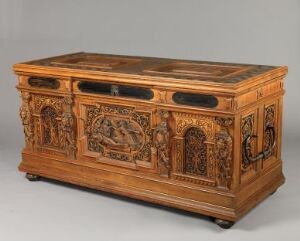  An elaborately carved wooden chest with detailed relief scenes on its front and sides, exhibiting a warm brown color with black accents on its flat top, placed against a neutral background. Artist name and title are unknown.