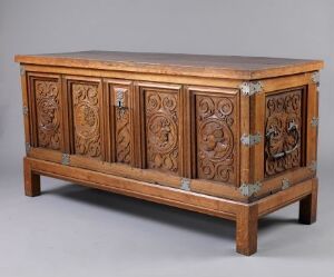  An antique wooden cabinet with a rectangular top, adorned with intricate carved panels featuring lion heads and foliate designs, set against a light gray background.