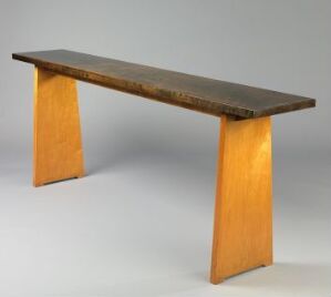  A minimalist wooden table with a dark brown tabletop and two vertical honey-colored legs, set against a plain background. Artist name and title are unknown.