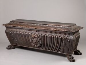  An intricately carved and varnished walnut chest titled "Kiste" by an unidentified artist, standing on paw-shaped feet with a fluted design on the sides and scalloped detailing along the rim and lid.