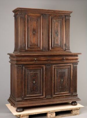  Traditional two-section wooden cabinet on a pallet, featuring raised, beveled panel doors with keyholes, and detailed trim, set against a neutral grey background. The upper section sits on the wider lower section, and the cabinet is made of dark brown wood with lighter brown accents.