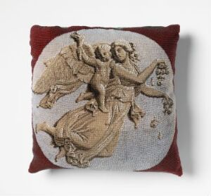  A decorative pillow with a classical embroidery of a winged figure in motion, rendered in beige and taupe on a light background, framed by a deep red border.