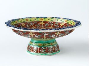  A handcrafted Bencharong porcelain bowl with a wide rim and pedestal base, intricately hand-painted with vibrant patterns of blue, green, red, and gold on a dark background. Artist unknown.