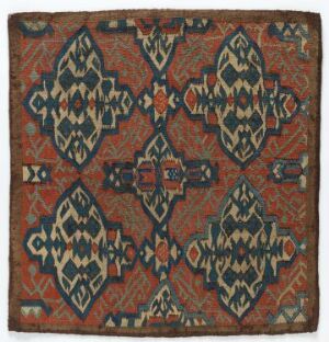  An intricately designed square textile with a symmetrical pattern of elongated diamond and triangular shapes in blue, white, and black, set against a red-brown background with red, green, and yellow accents, forming a large diamond shape, surrounded by a patterned border.