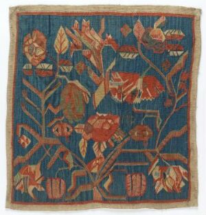  A square textile piece with a deep blue background and an intricate red, orange, and green botanical design, with leaves and blossoms intermingled, suggesting a fabric of historical or cultural significance. The edges are slightly frayed, implying age or use.