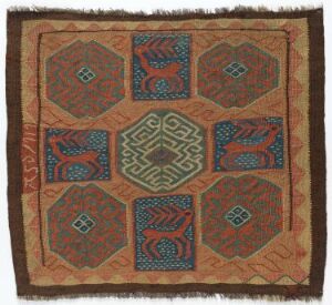  An intricately patterned handwoven textile or rug featuring a central maze-like octagonal motif in green and blue, surrounded by six smaller similar motifs, with a brown, orange, and terracotta base, bordered by a repeated geometric pattern in teal blue on a red background, finished with fringes. Artist name and title unknown.