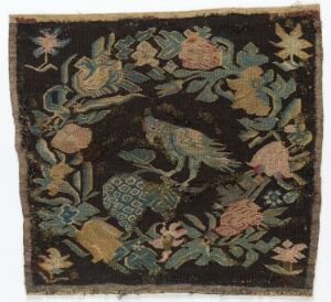  An antique textile fragment featuring an embroidered or woven garden scene with pineapples, pomegranates, assorted flowers, leaves, and two birds on a dark background, exhibiting a vibrant yet aged appearance.