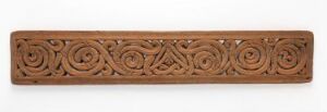  A narrow wooden panel intricately carved with symmetrical patterns of interlocking spirals and knots, set against an off-white background. The wood has a rich reddish-brown hue, and the carvings are deep enough to cast subtle shadows, emphasizing the detailed craftsmanship.