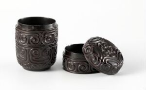  Two dark, intricately patterned cylindrical containers on a light background, one standing upright and the other lying on its side with the lid off, reflective of careful craftsmanship.