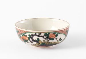  A decorative bowl with a glazed ivory interior and an ornate exterior pattern featuring green foliage and colorful florals below a salmon pink rim with delicate design work, set against a light grey background.