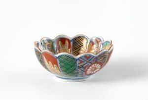  A small, colorful ceramic bowl with a wavy, flower-like rim, intricately patterned with red, green, orange, blue, and gold designs on a white background, suggesting a hand-painted, folk art style.