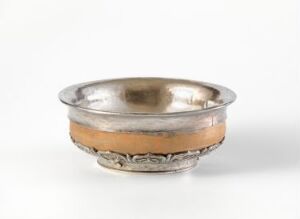  A shallow metallic bowl, possibly made of silver and gold, with an ornamental band, set against a plain, neutral background.