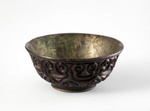  A dark bronze bowl with a green and gold patina, featuring intricate swirling relief patterns on the exterior, set against an off-white background.