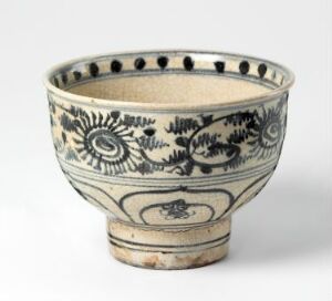  An old, handcrafted ceramic bowl with a footed base. It has a cream-colored background adorned with intricate indigo and black patterns, including sunburst floral motifs, geometric shapes, and a central oval emblem. The bowl shows signs of age. Artist name and title are unknown.