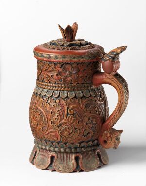  A richly decorated brown ceramic mug with floral embossments, a curved handle, and a lid topped with a small flower-shaped knob, set against a neutral background. Artist name and title are unknown.