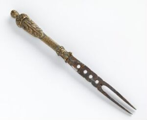  An antique two-pronged fork with a decorative metal handle featuring a floral or vine-like pattern in dark gold tones, and slender steel or silver prongs with circular holes, displayed against a light neutral background. The artist name and title are unknown.