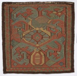  An antique rectangular textile with frayed edges, featuring a symmetrical geometric pattern with an olive-green background, burnt orange, warm beige, and slate gray designs, suggestive of traditional craftsmanship.