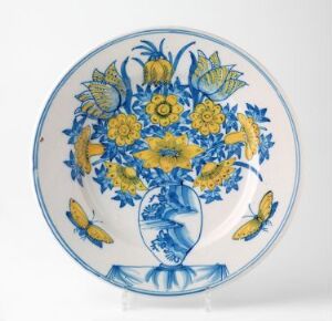  A round ceramic plate with an ornate design featuring a central bouquet of yellow flowers in a cobalt blue vase, surrounded by blue foliage and butterflies on a white background. The plate's rim is lined with a blue stripe and small blue decorative elements.