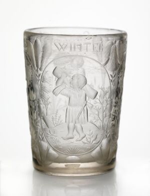  A cylindrical translucent glass with a tapering base, showcasing an etched design featuring a classical figure reminiscent of a cherub surrounded by stylized foliage or decorative motifs, set against a uniform gray background.