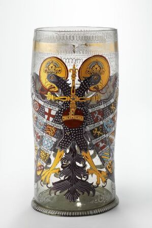  A traditional painted and engraved blown glass vessel featuring two birds with gold and black details facing each other across a shield with geometric and cross motifs, circled by detailed bands of color and decoration, against a light gray background. Artist unknown.