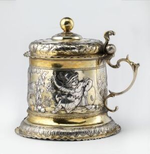  A decorative, metallic tankard with a lidded top, featuring golden and silver tones with embossed and engraved designs, set against a simple white background. Artist's name and title of the work are unknown.