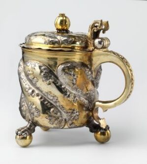  A silver drinking pitcher titled "Drikkekanne" by an unidentified artist, featuring embossed and gilded leaf-like patterns, a curved handle, a hinged lid with a golden knob, and is standing on three small spherical feet.