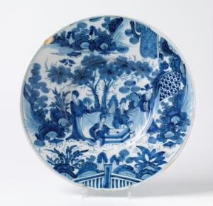  A ceramic plate with a detailed blue and white pastoral painting, featuring a peacock under a tree, floral patterns, and a fenced area, displayed against a white background. Artistname and title are unknown.