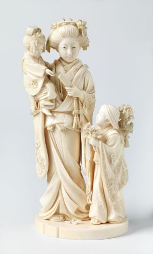  An ivory sculpture depicting the Virgin Mary holding baby Jesus, with a smaller figure holding a bouquet of flowers gazing towards them, set against a neutral grey background.