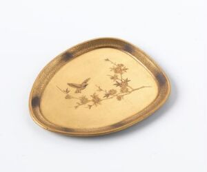  An oval-shaped metallic dish with a golden hue displays an embossed design of flowers and two birds in flight. The dish has a smooth rim and lies against a neutral off-white background, highlighting its warmth and ornate craftsmanship.