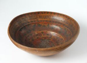  An aged bowl with a wide brim, displaying intricate patterns in muted orange and faded red on a dark brown background, suggestive of traditional craftsmanship.