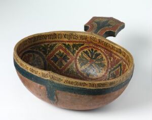  An ancient terracotta bowl with a broken handle, intricately decorated with geometric and floral patterns in green, yellow, orange, and brown on both the inside and outside, set against a neutral background.