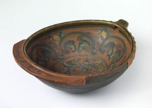  An ancient elliptical ceramic bowl with a single handle, exhibiting rustic ochre tones on the exterior and intricate, scroll-like motifs in dark hues on a burnt sienna glazed interior.