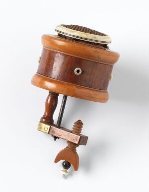  A vintage wooden and metal mechanical apparatus with a cylindrical body, a metal comb-like piece on top, and a screw clamp at the bottom for attaching to a surface. The polished brown wood and aged metallic elements suggest an old-fashioned device, possibly for sound generation or precise mechanical work. Artist name and title are unknown.