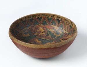  An ancient terracotta bowl with a plain exterior and a dark brown interior adorned with vibrant orange and yellow-orange patterns, including swirls, spirals, and stylized creatures. The rim features evenly spaced symbolic or script-like designs.