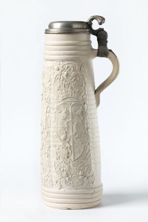  A traditional beer stein with intricate bas-relief designs and a pewter lid, set against a plain background, showcasing detailed craftsmanship and a muted color palette of off-white ceramic and silver-grey metal.