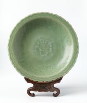  An antique green-glazed bowl with an embossed flower-like pattern in the center, resting on a carved wooden stand with scroll-like legs. Artistname and title remain unknown.