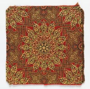  A piece of fabric or patterned paper featuring an intricate, symmetrical design with a central golden floral rosette on a deep red background, adorned with green and yellow patterns and unfinished edges. Artist name and title are unknown.