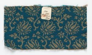  A rectangular swatch of fabric with a dark teal background and detailed golden-beige floral patterns, including a white paper tag with handwritten text attached to the upper right corner.