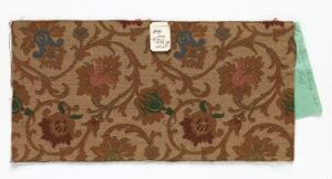  A rectangular swatch of heavy fabric with a brown background featuring a repeated pattern of darker brown ornamental designs and stylized greenish and reddish-brown floral motifs, centered by a white rectangular tag tied with a green string.