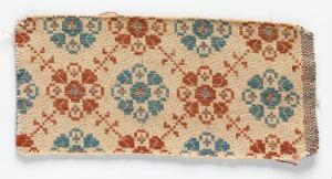  An image of a rectangular textile with a beige background featuring a symmetrical floral pattern in muted tones of rusty red, dusty blue, and sage green, with slightly frayed edges on one end. Artist name and title are unknown.