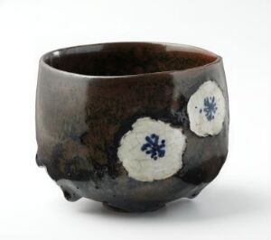  A handcrafted ceramic bowl with a deep brown color and two white, flower-emblazoned patches is displayed against a plain white background. The bowl's rim is slightly asymmetrical, adding to its artisanal charm.