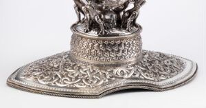  A silver-colored metal decorative object with elaborate etched details and ornate patterns on a flat, beveled base, featuring a pedestal-like upper section with less defined embellishments, set against a light background.