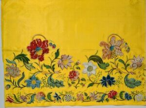  An embroidered floral pattern featuring a variety of colorful flowers and leaves on a golden yellow fabric. The design includes a prominent red flower with blue and yellow accents on the left, and a tapestry of pastel pinks, creams, lilacs, and greens to the right, all elaborately decorated with intricate stitching on a warm yellow background.