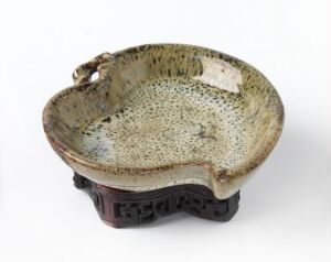  A handcrafted ceramic bowl with a speckled olive green and beige interior and a wavy rim, resting on an ornate dark wooden stand with traditional carved motifs.